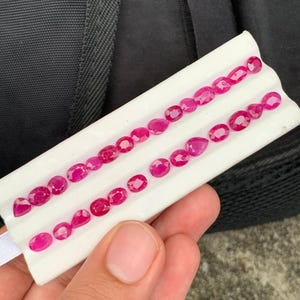 May include: A collection of small, faceted, oval and pear-shaped rubies arranged in two rows on a white display card. The gemstones exhibit a vibrant, deep pinkish-red hue with multiple facets that reflect light, showcasing their polished surfaces.