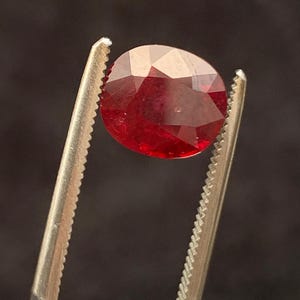 May include: A faceted oval-shaped red gemstone held by a pair of metal tweezers. The stone displays a deep, vibrant crimson hue with multiple geometric facets that reflect light across its polished surface, showcasing a clear and transparent quality.