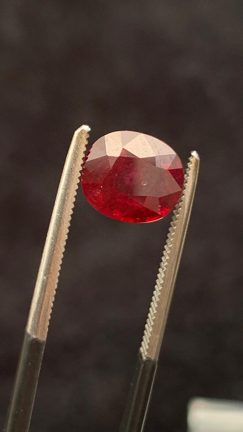 May include: A faceted oval-shaped red gemstone held by a pair of metal tweezers. The stone displays a deep, vibrant crimson hue with multiple geometric facets that reflect light across its polished surface, showcasing a clear and transparent quality.