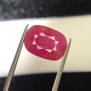 Puede incluir: Una piedra preciosa de color rojo rosado vibrante y corte cojín sostenida por pinzas de metal. La piedra presenta una inclusión o patrón blanco distintivo en forma de estrella de ocho puntas centrado en su superficie translúcida y pulida.