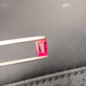 May include: A vibrant, rectangular-cut, deep pink gemstone held securely by a pair of silver-toned metal tweezers. The stone features a faceted surface that reflects light, showcasing its clear, saturated color and precise, sharp-edged geometric shape.