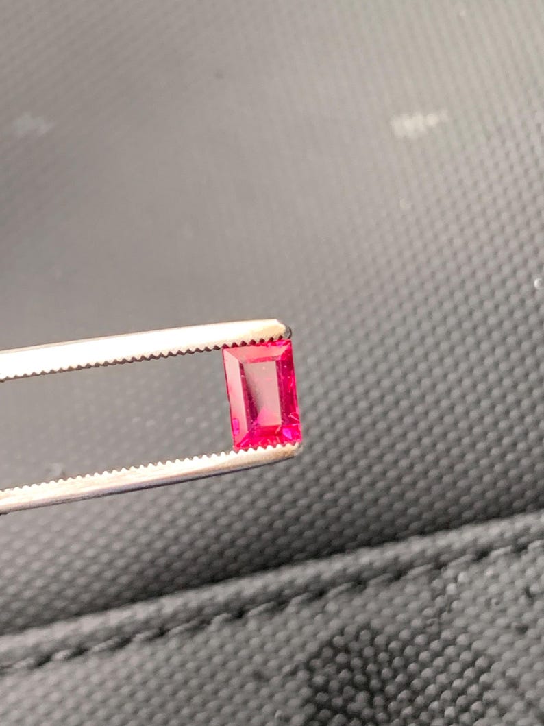 May include: A vibrant, rectangular-cut, deep pink gemstone held securely by a pair of silver-toned metal tweezers. The stone features a faceted surface that reflects light, showcasing its clear, saturated color and precise, sharp-edged geometric shape.