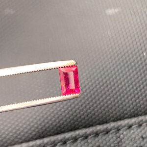 May include: A vibrant, rectangular-cut ruby gemstone with a deep pinkish-red hue, held securely by a pair of silver-toned metal tweezers. The gemstone features a faceted surface that reflects light, showcasing its clarity and saturated color profile.