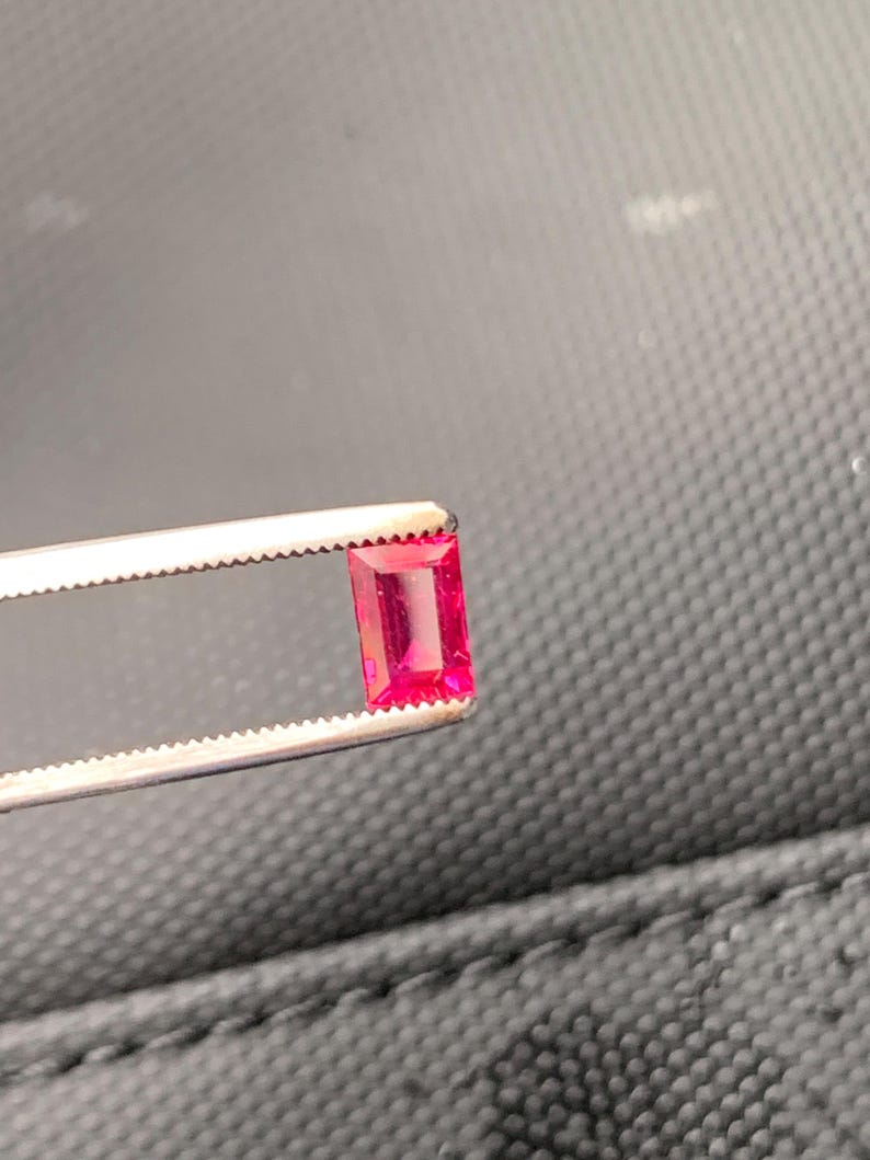 May include: A vibrant, rectangular-cut ruby gemstone with a deep pinkish-red hue, held securely by a pair of silver-toned metal tweezers. The gemstone features a faceted surface that reflects light, showcasing its clarity and saturated color profile.