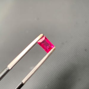 May include: A vibrant, rectangular-cut gemstone with a deep pinkish-red hue held by a pair of silver-toned metal tweezers. The stone features a faceted surface that reflects light, showcasing its transparent quality and precise geometric shape.