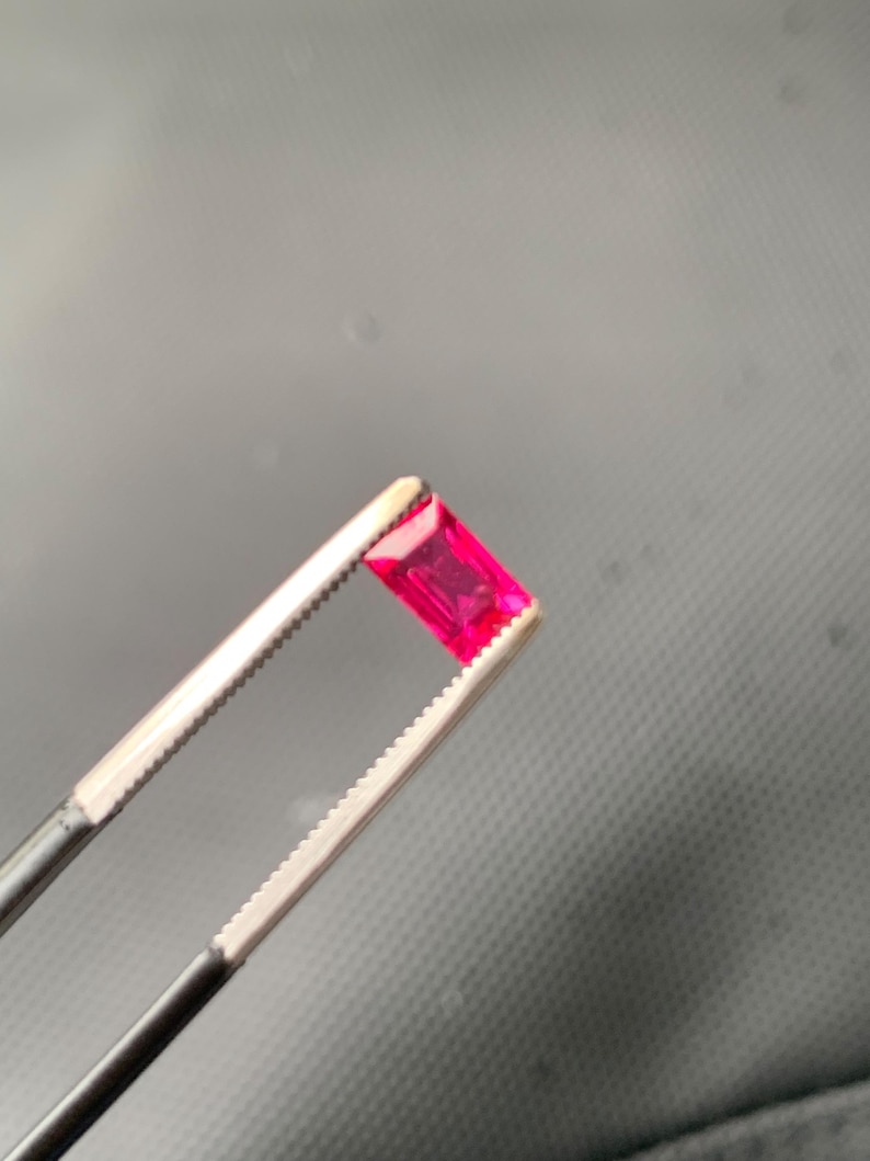 May include: A vibrant, rectangular-cut gemstone with a deep pinkish-red hue held by a pair of silver-toned metal tweezers. The stone features a faceted surface that reflects light, showcasing its transparent quality and precise geometric shape.