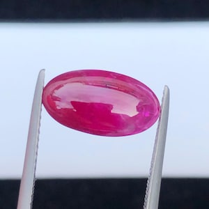 May include: A vibrant, oval-shaped, cabochon-cut gemstone with a deep magenta-pink hue. The stone features a smooth, polished surface that reflects light, held securely by a pair of silver-toned metal tweezers against a blurred background.