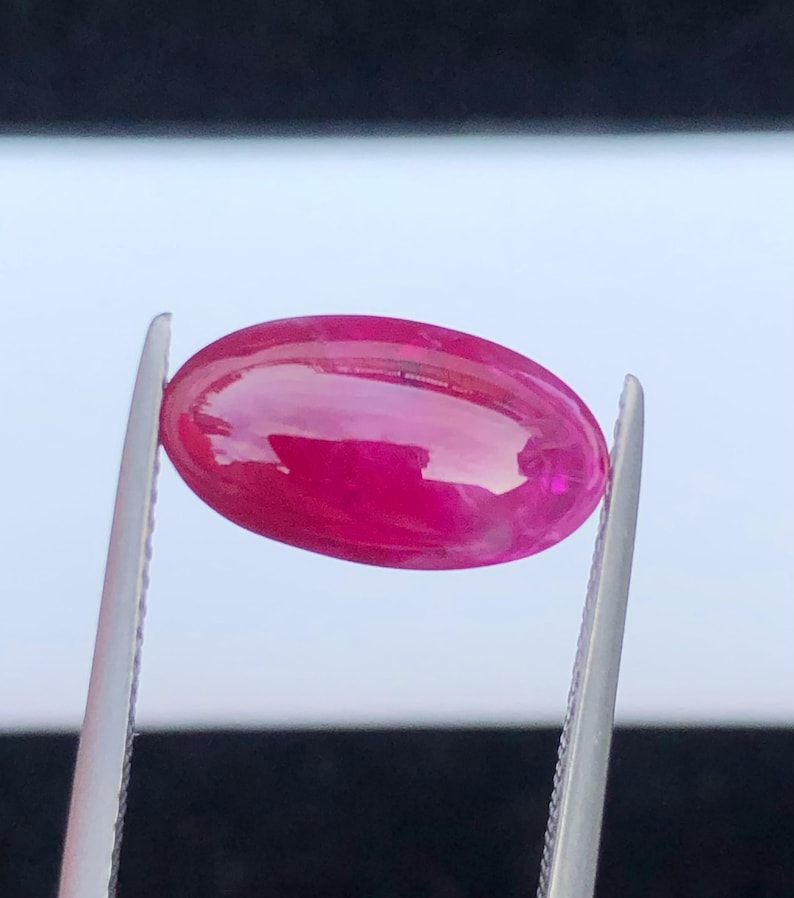 May include: A vibrant, oval-shaped, cabochon-cut gemstone with a deep magenta-pink hue. The stone features a smooth, polished surface that reflects light, held securely by a pair of silver-toned metal tweezers against a blurred background.