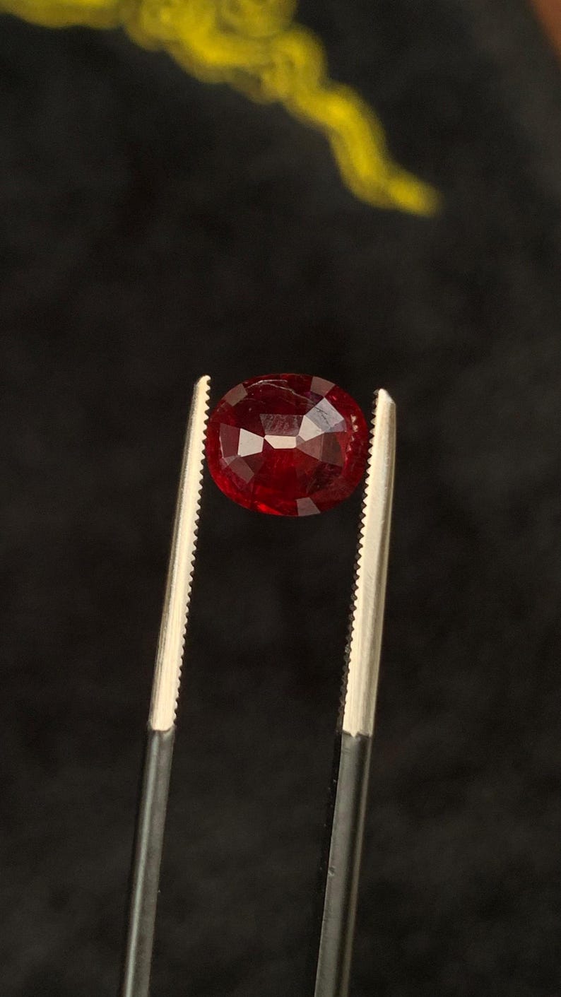 May include: A deep red, oval-cut gemstone with multiple facets reflecting light, held securely between the serrated metal tips of a pair of tweezers. The stone displays a rich, saturated crimson hue and a polished, reflective surface throughout.