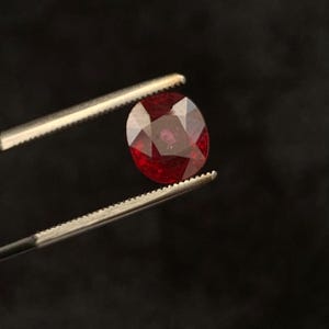 May include: A faceted, deep red, oval-shaped gemstone held by a pair of silver-toned metal tweezers. The stone exhibits a brilliant, polished surface with multiple geometric facets that reflect light, showcasing a rich, saturated crimson colour.
