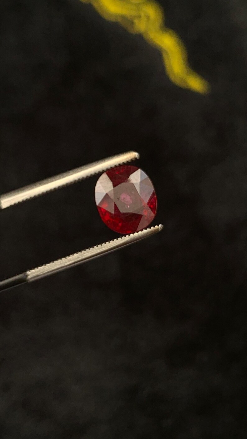 May include: A faceted, deep red, oval-shaped gemstone held by a pair of silver-toned metal tweezers. The stone exhibits a brilliant, polished surface with multiple geometric facets that reflect light, showcasing a rich, saturated crimson colour.