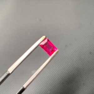 May include: A vibrant, rectangular-cut gemstone with a deep pinkish-red hue, held securely by a pair of metal tweezers. The stone features a faceted surface that reflects light, showcasing its clear, transparent quality and precise geometric shape.
