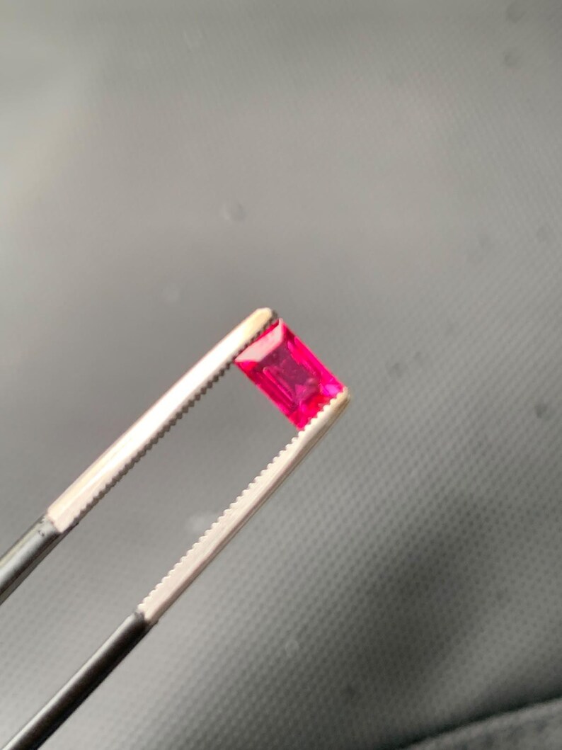 May include: A vibrant, rectangular-cut gemstone with a deep pinkish-red hue, held securely by a pair of metal tweezers. The stone features a faceted surface that reflects light, showcasing its clear, transparent quality and precise geometric shape.