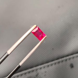 May include: A vibrant, rectangular-cut gemstone with a deep, saturated red color. The stone is held securely by a pair of metal tweezers with serrated tips, showcasing its faceted surface and translucent quality under bright lighting conditions.