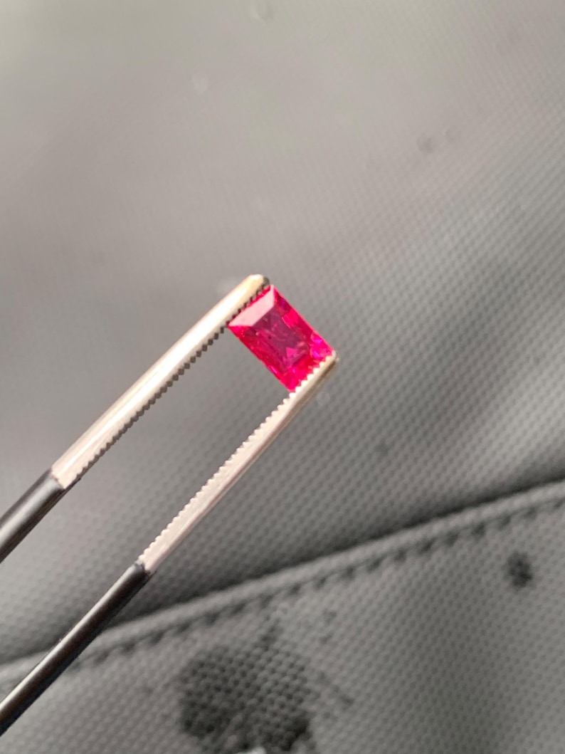 May include: A vibrant, rectangular-cut gemstone with a deep, saturated red color. The stone is held securely by a pair of metal tweezers with serrated tips, showcasing its faceted surface and translucent quality under bright lighting conditions.