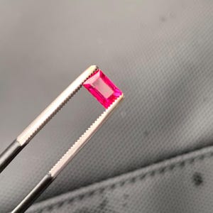 May include: A vibrant, rectangular-cut gemstone with a deep pinkish-red hue. The stone features a faceted surface that reflects light, showcasing its clarity and color. It is held securely by a pair of metal tweezers with serrated tips.