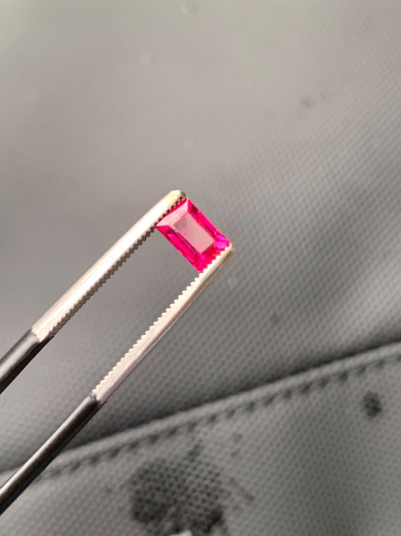 May include: A vibrant, rectangular-cut gemstone with a deep pinkish-red hue. The stone features a faceted surface that reflects light, showcasing its clarity and color. It is held securely by a pair of metal tweezers with serrated tips.