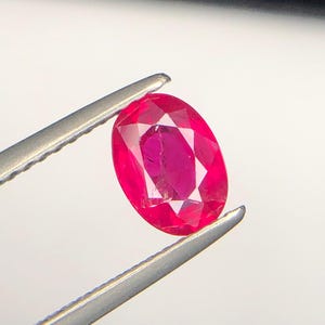 Peut inclure: Une pierre pr&eacute;cieuse rubis de taille ovale d'une teinte rouge vif avec une surface &agrave; facettes multiples, maintenue fermement par une pince en m&eacute;tal argent&eacute;. La pierre pr&eacute;sente une clart&eacute; interne et une r&eacute;flexion de la lumi&egrave;re sur ses facettes.