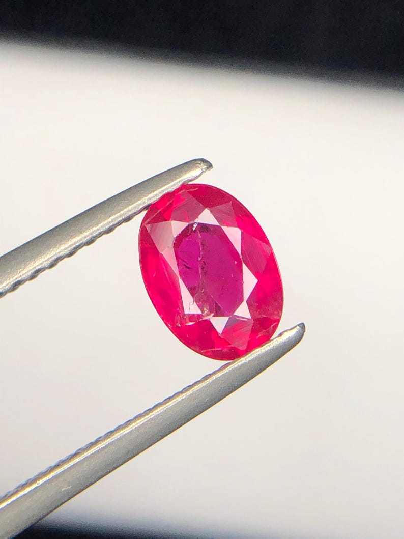 Peut inclure: Une pierre pr&eacute;cieuse rubis de taille ovale d'une teinte rouge vif avec une surface &agrave; facettes multiples, maintenue fermement par une pince en m&eacute;tal argent&eacute;. La pierre pr&eacute;sente une clart&eacute; interne et une r&eacute;flexion de la lumi&egrave;re sur ses facettes.