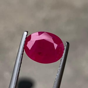May include: A vibrant, oval-cut gemstone with a deep, saturated magenta-red color. The stone features multiple triangular and kite-shaped facets across its surface, creating a reflective, polished appearance while held by metal tweezers.