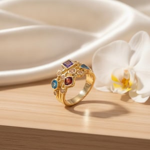 Peut inclure: Une bague en or ornée d'un groupe de pierres précieuses de formes variées, dont une pierre carrée violette, une pierre carrée rouge, une pierre ronde bleue et plusieurs petites pierres rondes transparentes sur un anneau en or texturé.