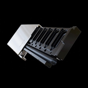 May include: A rectangular metallic device featuring a polished silver-toned top panel and a black base with a series of six elongated, oval-shaped recessed slots arranged in a row. The structure has a clean, industrial design with sharp edges.