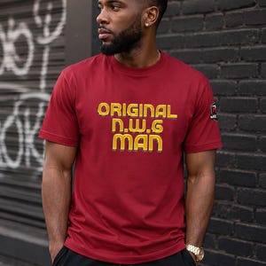 May include: A short-sleeved crew neck t-shirt in a deep maroon color. The front features the text "ORIGINAL n.w.g MAN" in bold, yellow, block-style lettering with a dark outline. A small black and red logo patch is visible on the left sleeve.