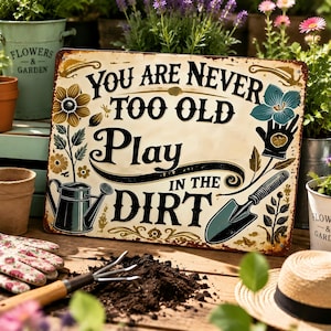 May include: A rectangular metal sign with a cream background and rust-colored edges. The text reads, "YOU ARE NEVER TOO OLD TO PLAY IN THE DIRT." The design features illustrations of a yellow flower, a blue flower, a watering can, and a garden trowel.