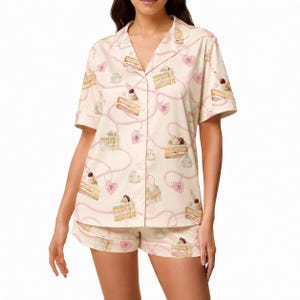 Puede incluir: Un conjunto de pijama color crema que consta de una camisa de manga corta con botones y pantalones cortos a juego. La tela está decorada con un patrón repetitivo de trozos de pastel, copetes de nata montada y dijes rosas en forma de corazón.
