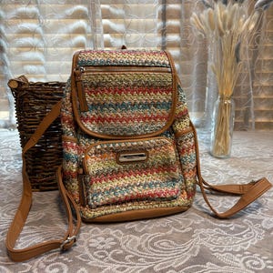 Puede incluir: Una mochila tejida multicolor con un patrón de chevrón en tonos rojo, azul, amarillo y beige. El bolso incluye ribetes de piel sintética color canela, correas ajustables, un compartimento principal con cremallera y un bolsillo frontal con placa plateada.