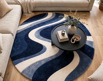 May include: A large circular area rug featuring a flowing, wave-like pattern in shades of deep navy blue, slate blue, and cream. The rug has a thick, textured pile and is positioned on a light-coloured wooden floor beneath a round black coffee table.