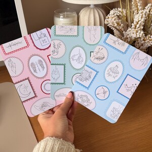 May include: Three rectangular cards featuring various line art illustrations enclosed in decorative frames. The cards are pink, light green, and light blue, displaying motifs like wings, a heart, a deer, a cauldron, and the text "light it up".