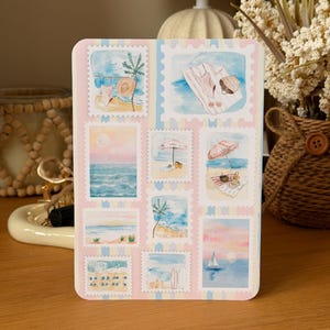 May include: A rectangular sticker sheet featuring ten individual postage stamp designs with watercolour illustrations of beach scenes, including palm trees, umbrellas, sailboats, sunsets, and coastal landscapes in soft pastel blue, pink, and tan tones.