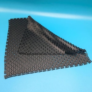 Può includere: Un tappetino nero flessibile caratterizzato da un motivo a griglia intrecciata ripetitivo composto da piccoli collegamenti quadrati. Il tappetino è ripiegato su un angolo, rivelando la struttura geometrica coerente del materiale scuro.