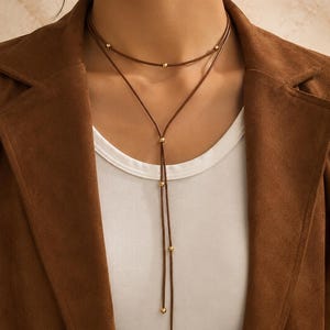 Puede incluir: Un collar de cordón marrón con un diseño de capas y pequeñas cuentas de metal dorado espaciadas a lo largo de las hebras. El collar incluye una sección larga de estilo lazo que cuelga por el centro, acentuada con cuentas doradas adicionales.
