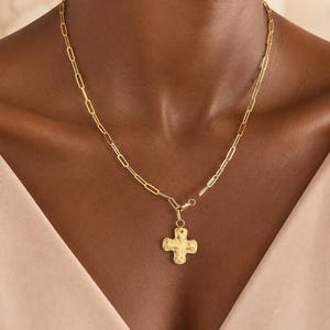Puede incluir: Un collar de tono dorado con un diseño de cadena de eslabones rectangulares tipo clip. El collar incluye un colgante central con forma de cruz con una superficie texturizada y martillada, suspendido de una pequeña anilla circular.
