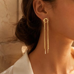 Peut inclure: Une boucle d'oreille pendante dorée composée d'un clou en forme de nœud circulaire relié à deux longues chaînes serpent plates et fines qui pendent verticalement. La boucle d'oreille présente une finition métallique polie et un design épuré.