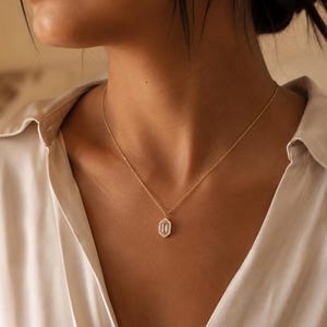 Puede incluir: Un collar de cadena dorada con un colgante central que presenta una piedra clara rectangular alargada rodeada por un halo de pequeñas piedras blancas. El colgante tiene una forma geométrica multifacetada y cuelga de una cadena metálica fina.