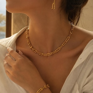 Könnte beinhalten: Ein goldfarbenes Schmuckset bestehend aus einer Halskette, einem Armband und einem Paar Ohrhänger. Jedes Stück weist ein wiederkehrendes U-förmiges Glieddesign mit kleinen kugelförmigen Akzenten an der Basis jedes Glieds auf.