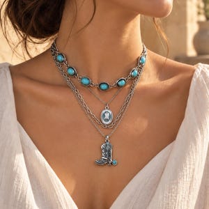 Puede incluir: Un conjunto de collares superpuestos en tono plateado que incluye una gargantilla con piedras turquesas ovaladas, una cadena intermedia con un colgante ovalado de centro azul claro y una cadena inferior con un dije de bota vaquera en plata.