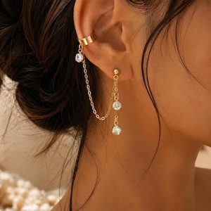 Puede incluir: Un pendiente tipo ear cuff dorado conectado por una cadena fina a un pendiente de botón. El ear cuff presenta un cristal redondo transparente, mientras que el pendiente de botón suspende dos cristales redondos transparentes adicionales.