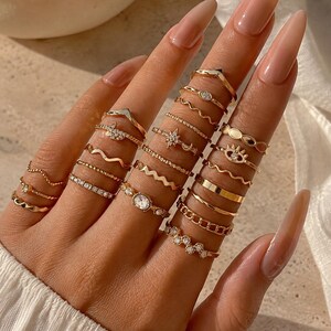 Puede incluir: Una colección de anillos dorados usados en varios dedos, con diversos diseños que incluyen bandas en forma de chevron, bandas onduladas finas, texturas de cuentas, un motivo de estrella, un dije en forma de ojo y pequeñas piedras claras.