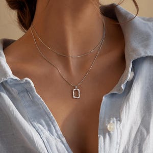 Puede incluir: Un collar de plata en capas con dos cadenas delicadas. La cadena más corta es un hilo fino y sencillo, mientras que la más larga sostiene un pequeño colgante rectangular con una superficie texturizada e irregular, sobre una camisa azul claro.