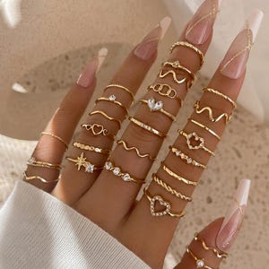 Könnte beinhalten: Eine Sammlung goldfarbener Ringe an den Fingern mit verschiedenen Designs, darunter dünne Bänder, Wellenmuster, Herzformen, Sternmotive und kleine klare Steine. Die Ringe sind in einer geschichteten Anordnung über mehrere Finger verteilt.