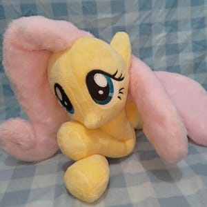 May include: A plush toy of a yellow pony with a long, soft pink mane and tail. The pony has large, embroidered teal eyes with black pupils and long eyelashes. The body is made of a soft, fuzzy yellow fabric, and the mane and tail are made of a plush, light pink material.