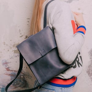 May include: A dark blue leather backpack with a rectangular flap closure, a single vertical strap detail, and thin adjustable shoulder straps. The bag features visible stitching along the edges and a smooth matte finish on the leather surface.