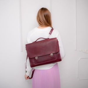 May include: A deep burgundy leather satchel bag with a top handle and a single adjustable shoulder strap. The bag features a large front flap secured by two silver-toned metal push-lock closures, providing a structured and functional design.