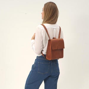 May include: A medium-sized, structured brown leather backpack featuring a rectangular main compartment with a fold-over flap closure, a single central strap fastener, and two slim shoulder straps, worn on the back over a white shirt and blue jeans.