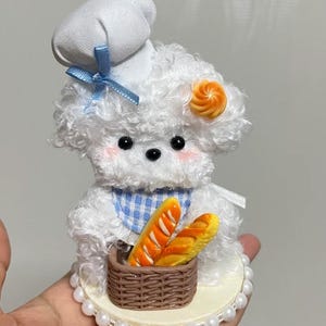 May include: A small white plush dog figurine wearing a white chef hat with a blue ribbon, a blue and white chequered bib, and a small orange swirl accessory. The dog holds a brown woven basket containing two miniature golden-brown baguette loaves.