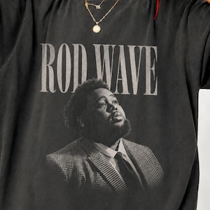 May include: A charcoal gray short-sleeve t-shirt featuring a vintage-style graphic print. The design includes the text ROD WAVE in a bold serif font above a black and white portrait of a person wearing a suit jacket and a collared shirt.